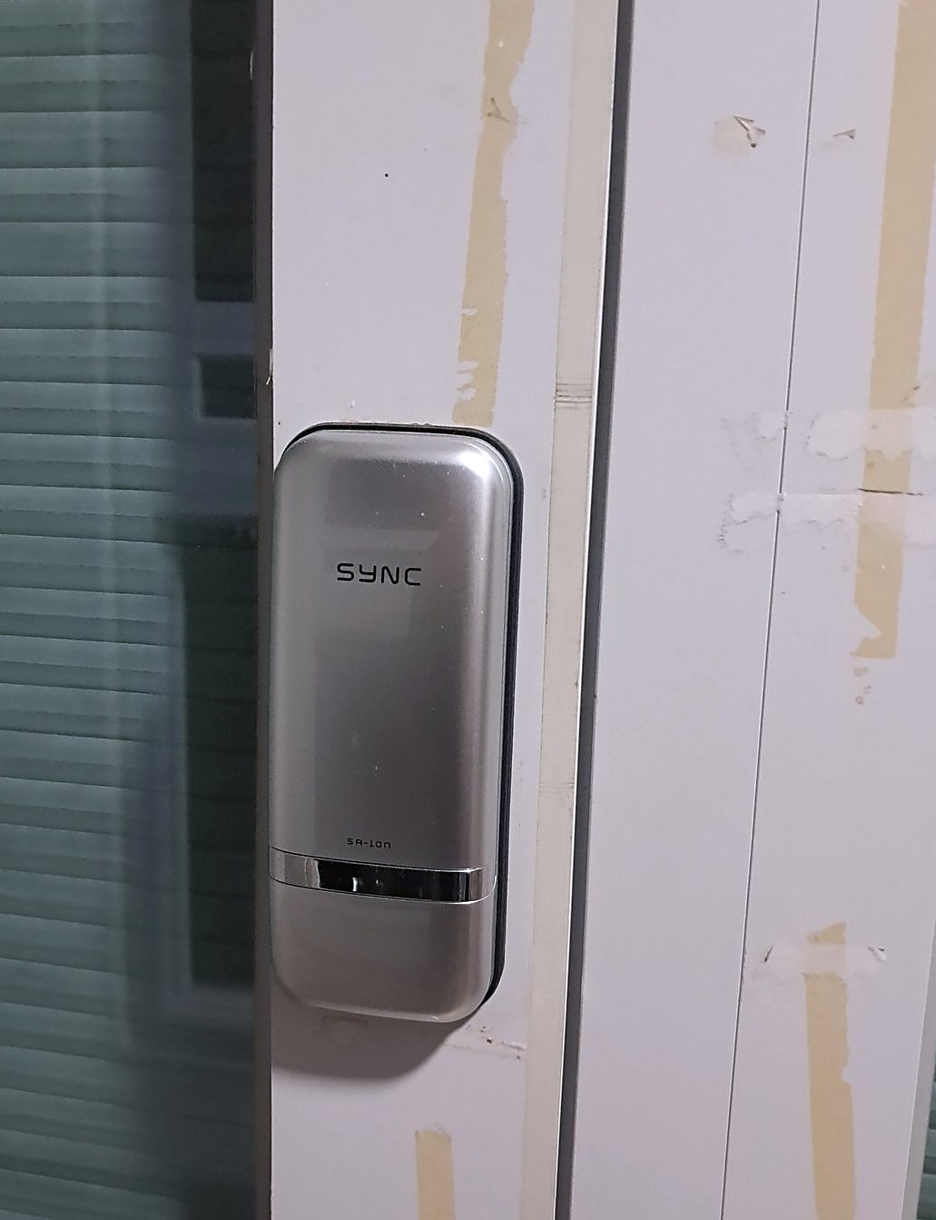 시흥시 연성동 출장 — 반투명 유리 옆 흰 문틀에 붙은 은색 도어락 본체와 문에 붙은 베이지 테이프 조각